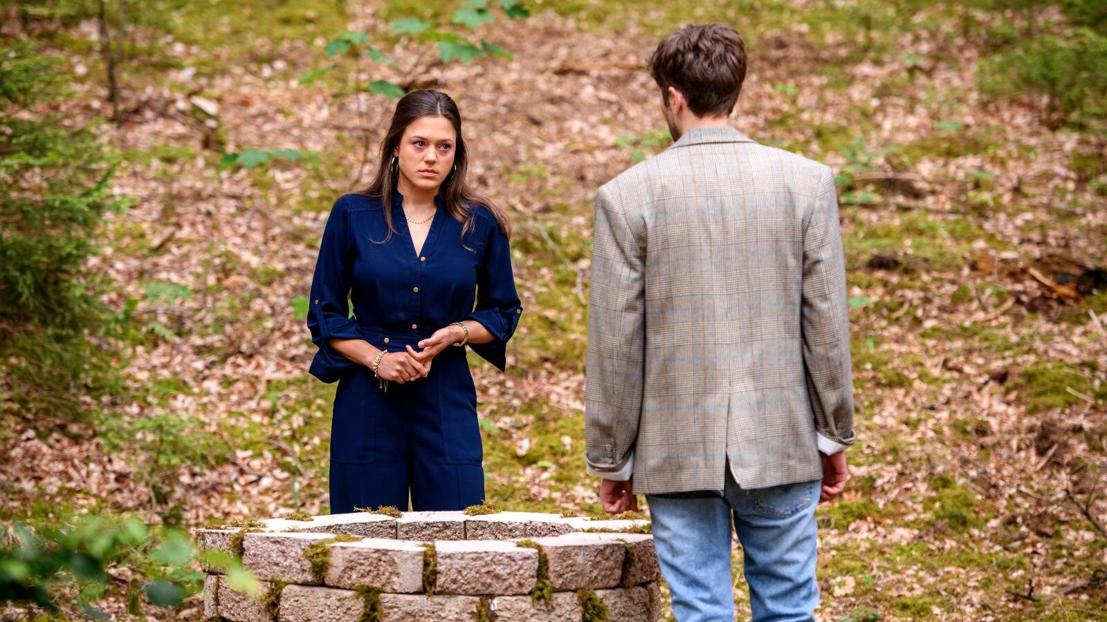 Eine junge Frau in dunkelblauem Kleid steht mit besorgtem Blick vor einem mit Moos bewachsenen Brunnen im Wald, ein Mann mit Jackett ihr gegenüber. | © ARD/WDR/Christof Arnold