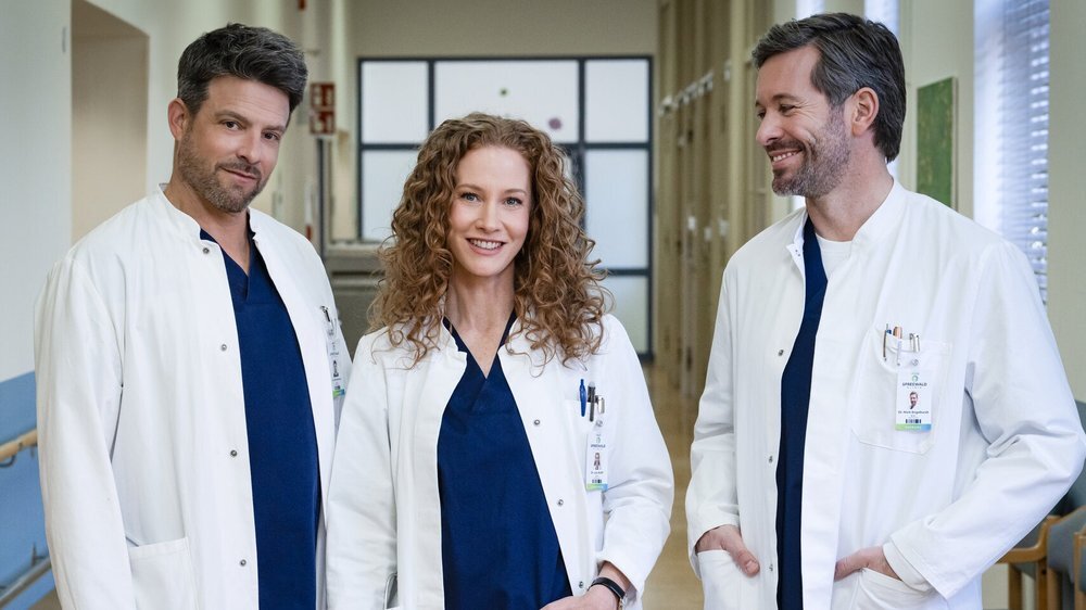 Eine rothaarige Ärztin mit Locken steht zwischen zwei männlichen Kollegen in einem hellen Krankenhausgang, alle tragen Namensschilder. | © Sat.1/Joyn/Claudius Pflug