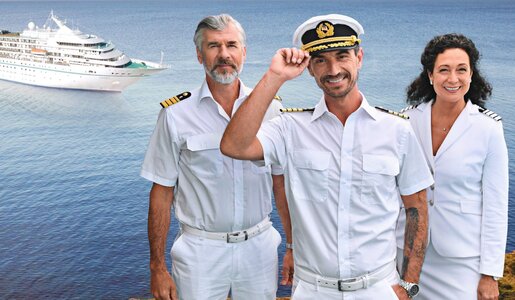 Drei Crewmitglieder in weißer Kapitänsuniform stehen lächelnd vor einem Kreuzfahrtschiff auf blauem Meer, einer hebt die Mütze. | © ZDF / Dirk Bartling