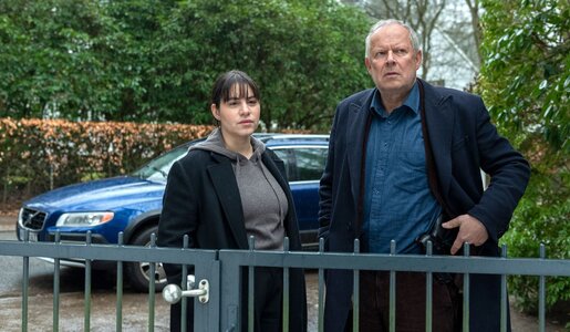 Eine junge Frau mit dunklen Haaren und ein älterer Mann mit ernstem Blick stehen vor einem Metalltor, hinter ihnen ein blauer Volvo und Büsche. | © NDR/Thorsten Jander