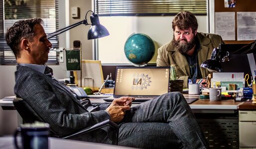 Ein ernster Mann im Anzug sitzt mit Handy in der Hand, während ein bärtiger Kollege ihn am Schreibtisch nachdenklich mustert. | © RTL / Behrus Yazdanfar / Redseven Entertainment