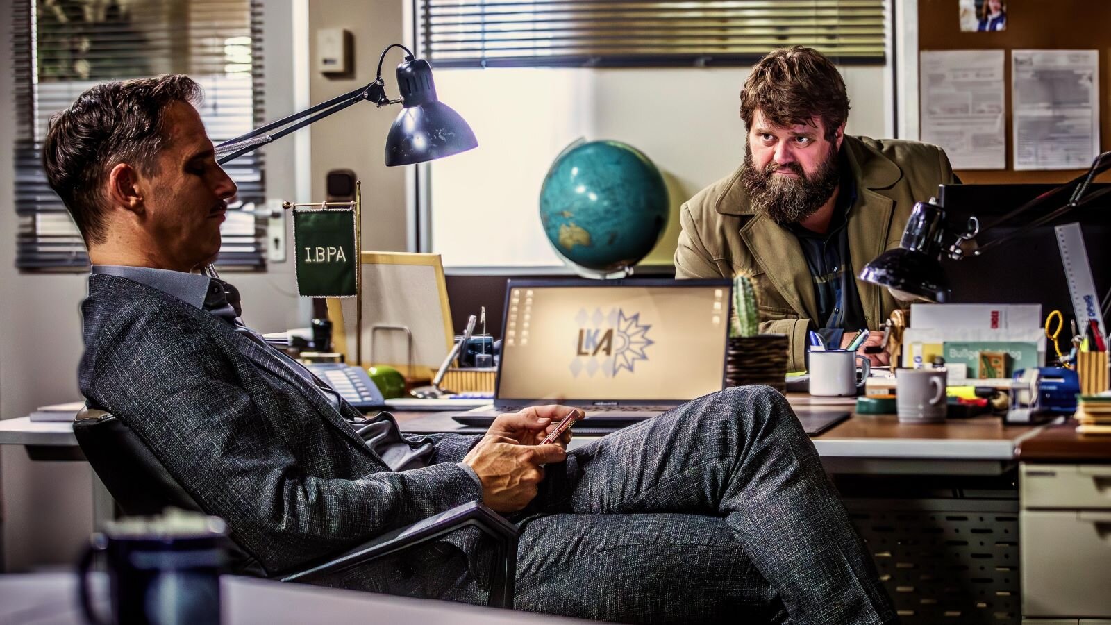 Ein ernster Mann im Anzug sitzt mit Handy in der Hand, während ein bärtiger Kollege ihn am Schreibtisch nachdenklich mustert. | © RTL / Behrus Yazdanfar / Redseven Entertainment