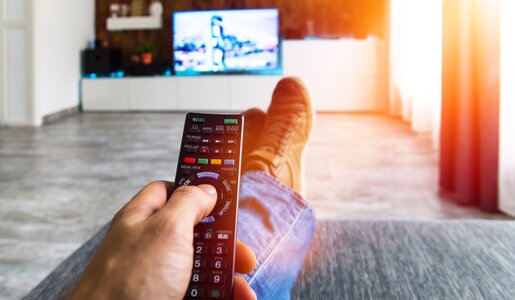 Mann sitzt auf Couch mit Fernbedienung in der Hand und vorne an Wand Fernseher an | © Adobe Stock/ghazii