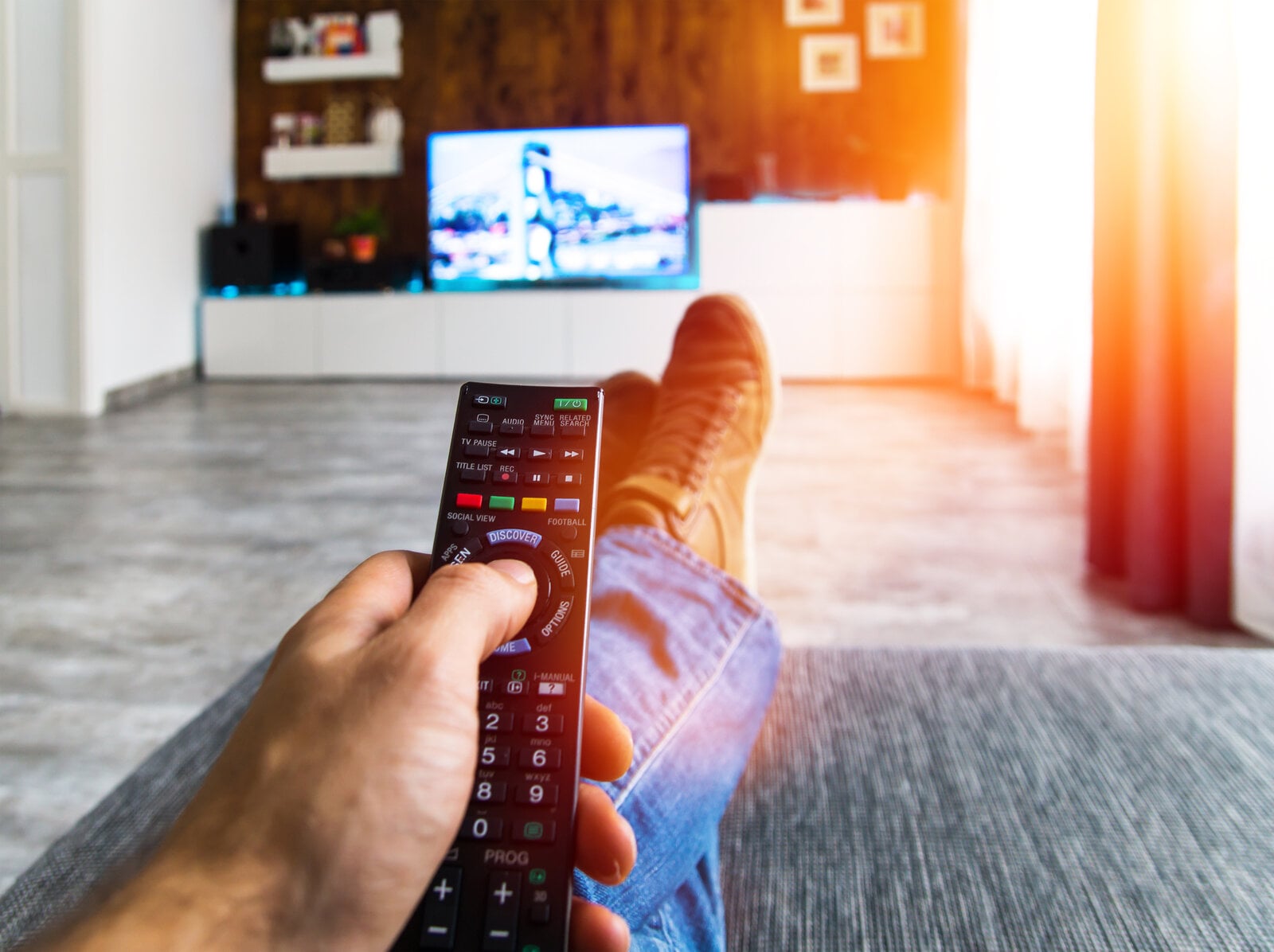 Mann sitzt auf Couch mit Fernbedienung in der Hand und vorne an Wand Fernseher an | © Adobe Stock/ghazii
