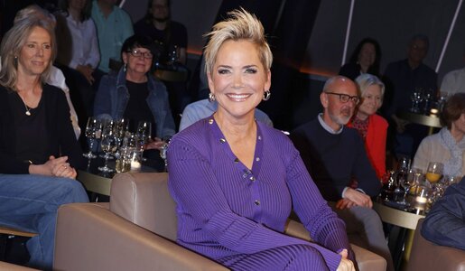 Strahlende Frau mit kurzem, blondem Haar in lilafarbenem Outfit sitzt vor einem Publikum an kleinen Tischen mit Weingläsern. | © IMAGO / Future Image / H. Hartmann