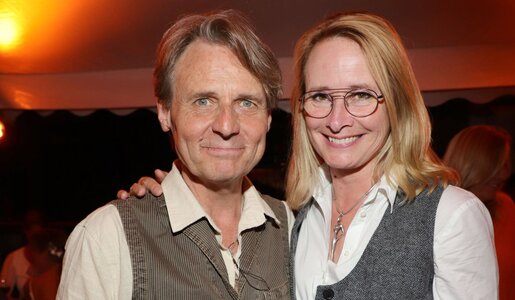 Eine blonde Frau mit Brille und ein grauhaariger Mann lächeln nebeneinander vor dunklem Hintergrund unter orangefarbenem Licht. | © IMAGO / Eventpress Becher