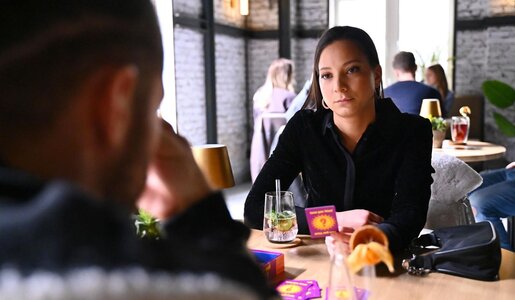 Zwei Menschen spielen an einem Holztisch ein Fragespiel in gemütlicher Bar, Frau blickt ruhig und konzentriert auf ihren Gegenüber. | © RTL / Julia Feldhagen
