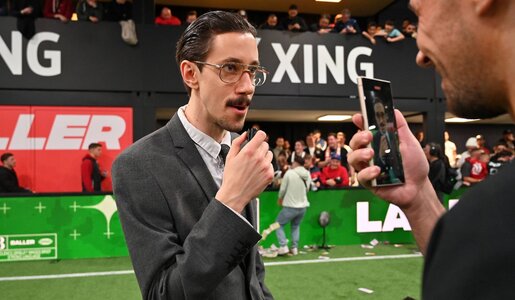 Junge Mann mit Schnurrbart und Brille im grauen Anzug spricht ernst in ein Mikrofon, während er gefilmt wird, Zuschauer im Hintergrund. | © IMAGO / Ulrich Hufnagel