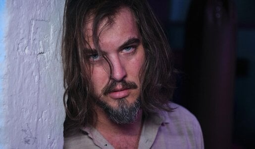 Ein junger Mann mit schulterlangem Haar und grauem Bart lehnt erschöpft mit starrem Blick an einer violett beleuchteten Wand. | © RTL / Stefan Behrens