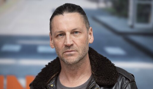 Ein ernster, mittelalter Mann mit zurückgebundenen dunklen Haaren trägt eine Lederjacke mit Fellkragen vor unscharfem Stadtmotiv. | © IMAGO / Sven Simon