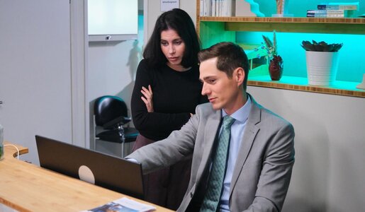 Ein elegant gekleideter Mann erklärt einer schwarzgekleideten Frau mit ernstem Blick etwas am Laptop in modernem Empfangsbereich. | © RTL / Stefan Behrens