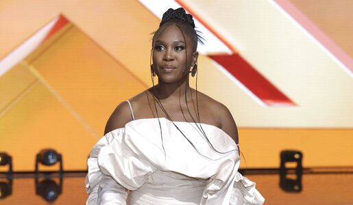 Elegante Frau in weißem Off-Shoulder-Kleid mit Puffärmeln steht mit ernstem Blick vor einer Bühne mit geometrischem Lichtdesign. | © IMAGO / Future Image / R. Schmiegelt