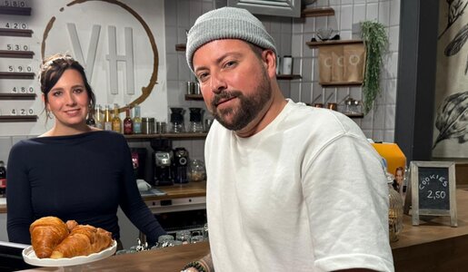 Ein bärtiger Mann mit grauer Mütze lehnt entspannt an einer Café-Theke, hinter ihm lächelt eine junge Frau in dunklem Oberteil. | © RTL / Frank Pick