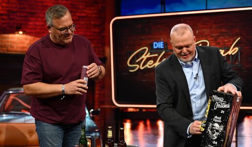 Zwei Männer öffnen lachend Bierdosen vor Adventskalender und Flaschen in TV-Studio mit Backsteinwand. | © RTL / Raab Entertainment / Julia Feldhagen