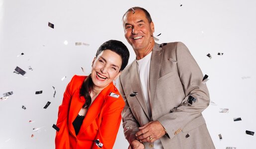 Ein fröhliches Paar mittleren Alters steht eng beieinander, sie im roten Blazer, er im beigen Anzug, Konfetti fällt vor hellem Hintergrund. | © RTL / Julia Feldhagen