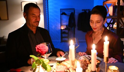 Ein stilvoll gekleidetes Paar blickt beim Abendessen auf den gedeckten Tisch mit Kerzen, Gläsern, Rose und kleinen Speisen. | © RTL / Julia Feldhagen