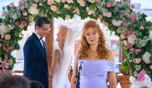 „My Ex - Die Liebenden“: Jan (Šimon Obdržálek) und Emma (Alli Neumann) stehen unter einem begrünten Traubogen mit weißen und rosa Rosen und schauen sich sehr verliebt an. Ivy (Palina Rojinski) steht seitlich vor ihnen vor einem Mikrophon und hält eine Rede. | © ZDF/Tomáš Mikule