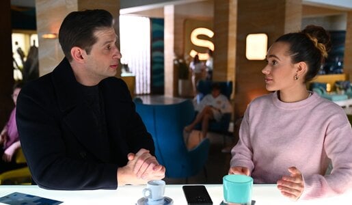 Junger Mann im schwarzen Mantel spricht mit junger Frau im rosa Pullover an hellem Tisch in moderner Lounge. | © RTL / Julia Feldhagen