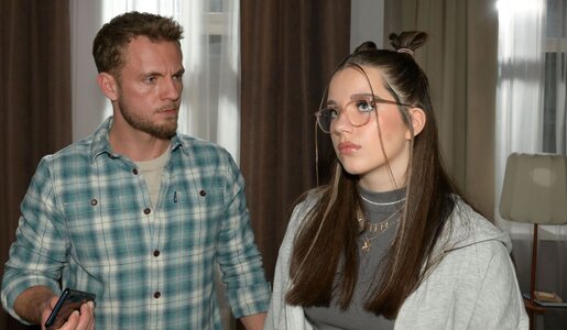 Ein bärtiger junger Mann hält ein Handy und schaut besorgt zu einer Teenagerin mit Brille vor dunklen Vorhängen. | © RTL / Rolf Baumgartner