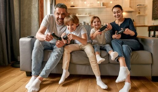Vater, Mutter und zwei Kinder sitzen fröhlich auf dem Sofa und halten Controller in der Hand.  | © Svitlana / stock.adobe.com