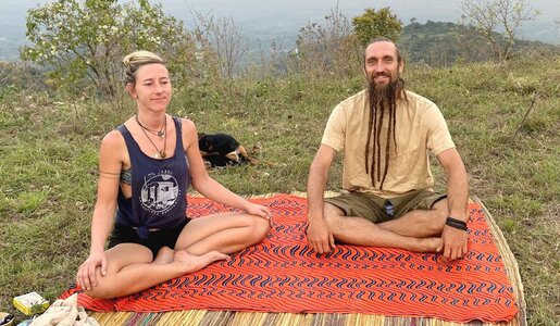 Eine blonde Frau und ein Mann mit langen Dreadlocks sitzen meditierend auf einer orangefarbenen Decke in einer grünen Hügellandschaft. | © RTL