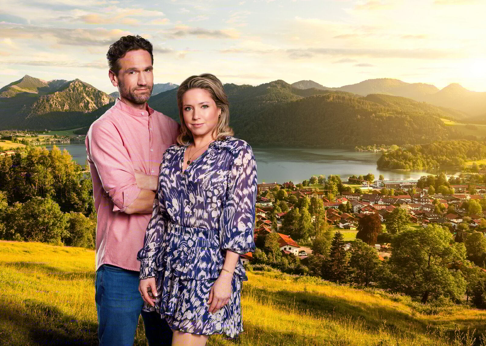 Ein Mann im rosafarbenen Hemd und eine Frau im blauen Kleid stehen vor einem sonnigen Alpenpanorama mit einem Dorf am See. | © Seven.One/Marc Rehbeck