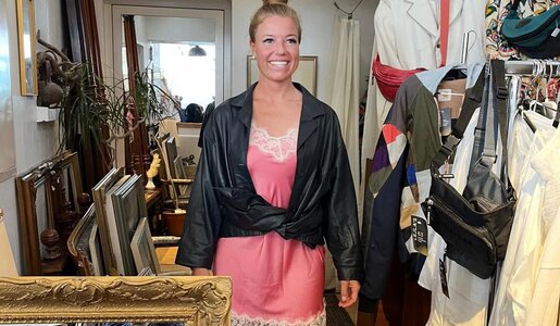 Blonde Frau mit hochgestecktem Haar trägt ein rosa Spitzenkleid und schwarze Lederjacke, umgeben von Vintage-Möbeln und Kleidern. | © RTL/Constantin Ent.