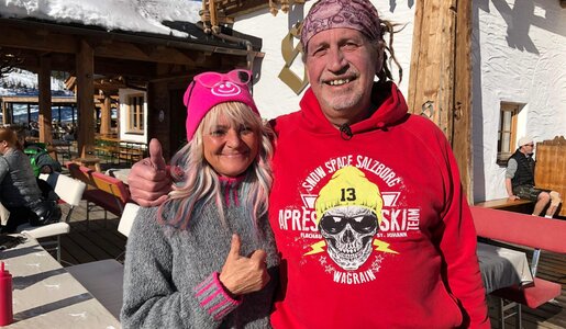 Ein Mann in rotem Hoodie und eine Frau mit rosa Mütze stehen bei Sonnenschein vor einer rustikalen Skihütte. | © RTL/99pro media
