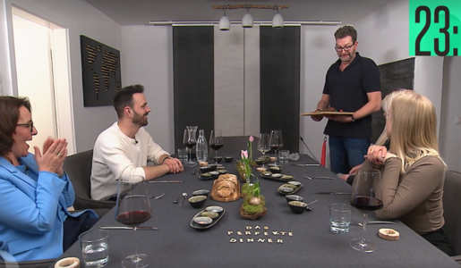 Der Gastgeber in dunkler Kleidung serviert lächelnd Speisen, während drei Gäste gespannt am gedeckten Tisch sitzen. | © Screenshot Vox/RTL+ 