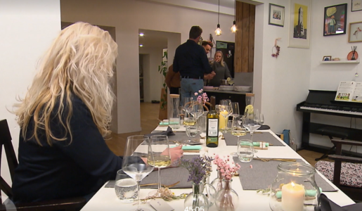 Eine blonde Frau sitzt an einem gedeckten Tisch, während im Hintergrund drei Personen in einer modernen Küche stehen. | © Screenshot Vox/RTL+ 