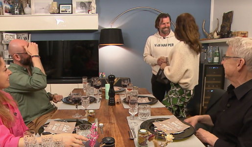 Ein Mann im weißen Hoodie schüttelt einer Frau in grünem Rock die Hand, während drei andere Personen am Esstisch sitzen und lachen. | © Screenshot Vox/RTL+ 