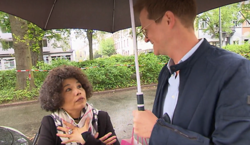Ein Mann und eine Frau stehen unter einem Regenschirm, sie spricht gestikulierend, im Hintergrund ein parkähnliches Stadtgebiet. | © Screenshot Vox/RTL+ 