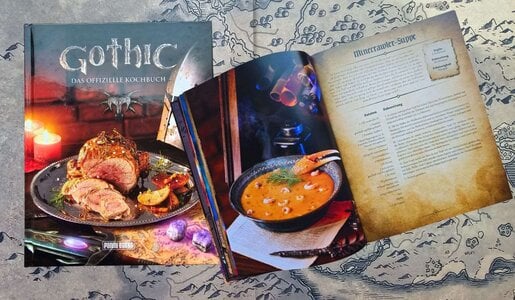 Offizielles "Gothic"-Kochbuch mit rustikaler Fleischplatte und dampfender Krabbensuppe, auf einer Landkarte liegend. | © Sabrina Forst